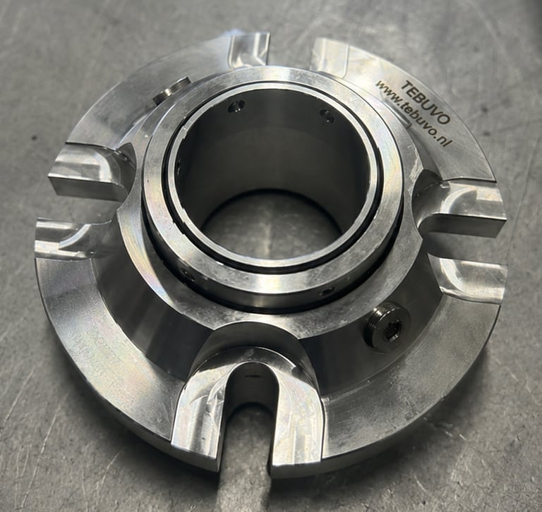 Close-up of a metallic mechanical pump seal with central hollow bore and curved engagement slot on gray surface
